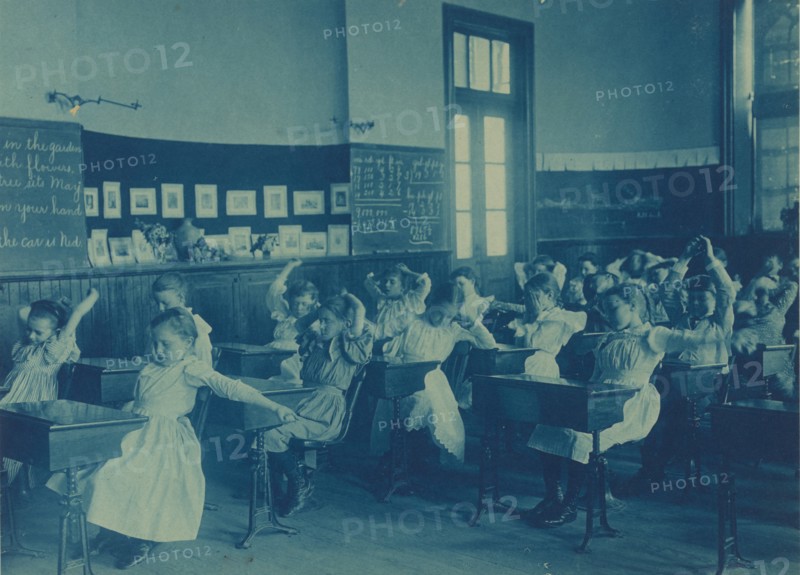 Children doing calisthenics while sitting at their desks in a classroom, 5th Division public schools, Washington, D.C., (1899?).