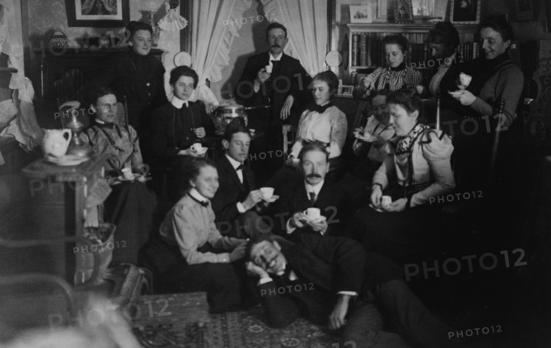 Group portrait of friends at social gathering, seated and standing with tea cups, in domestic setting, between 1890 and 1910.
