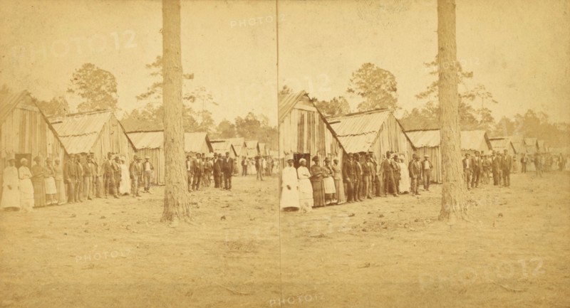 Group lined up in front of a row of shacks, (1868-1900?). Creator: Unknown.