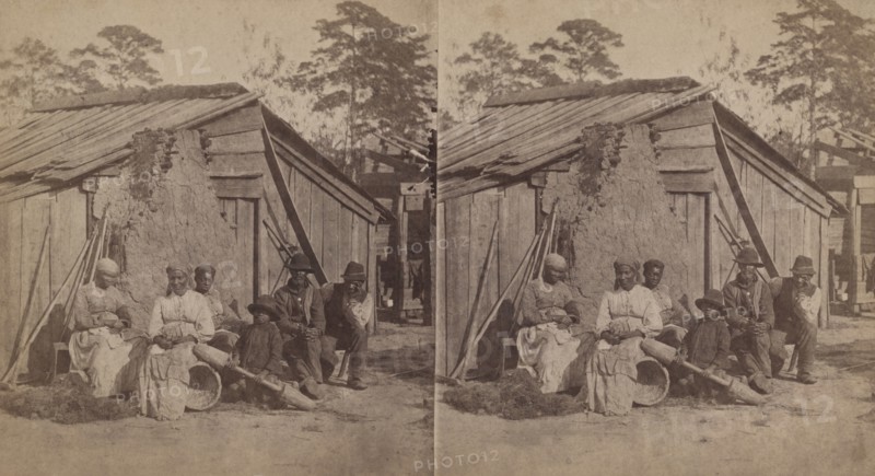 Family group sitting in front of a wooden shack, (1868-1900?). Creator: Unknown.
