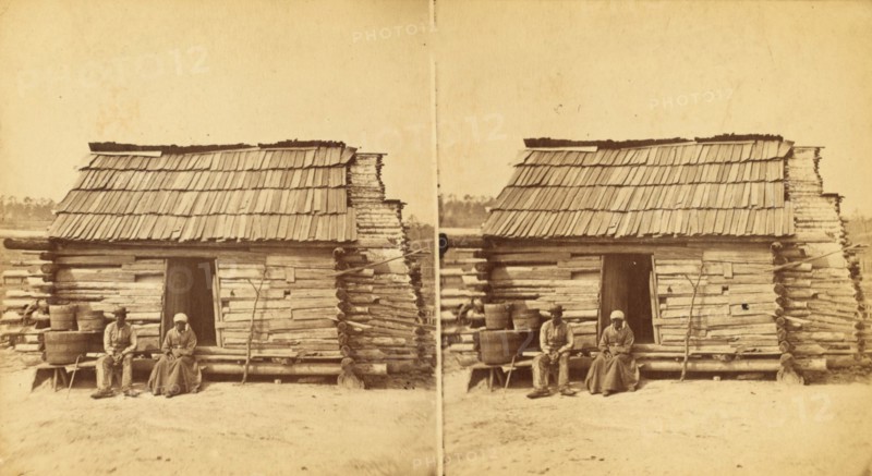 Uncle Ben's cabin, [Elderly couple sitting in front of a cabin], (1868-1900?). Creator: Unknown.