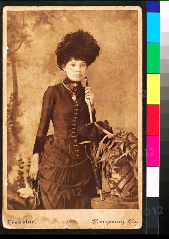 Standing woman, wearing dress with bustle, holding purse at side and umbrella resting..., 1886. Creator: Unknown.