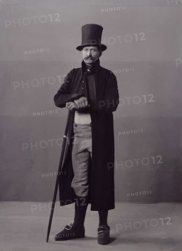 A man poses in folk costume with a long coat and a top hat, 1880-1907. Creator: Helene Edlund.