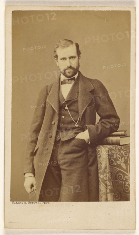 Unidentified well-dressed bearded man standing, elbow on books on a table, 1865-1870. Creator: Bayard & Bertall.