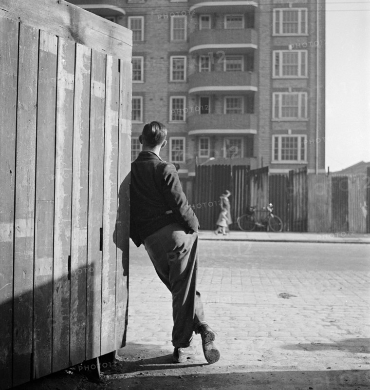 A man leaning on a street corner hoarding looks across the road to where a woman and child pass corrugated iron sheets enclosing an apartment block, possibly Rugmere on Ferdinand Street, Camden, 1945-1950.