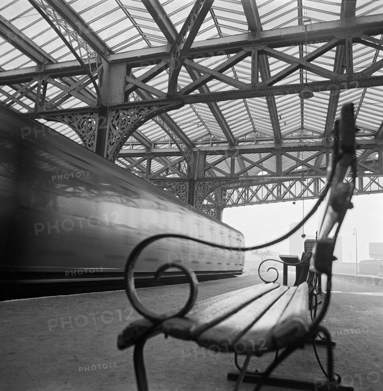 Looking along a bench with curved wrought iron arms on a platform at Waterloo Station with a train passing at speed in the background, 1960-1972.