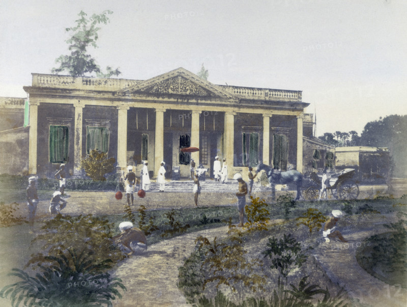 Residence of a European, India, 1850. Hand-coloured photograph showing a 'sahib' reclining on his veranda, while Indians tend to horses, carry water, and weed the garden.
