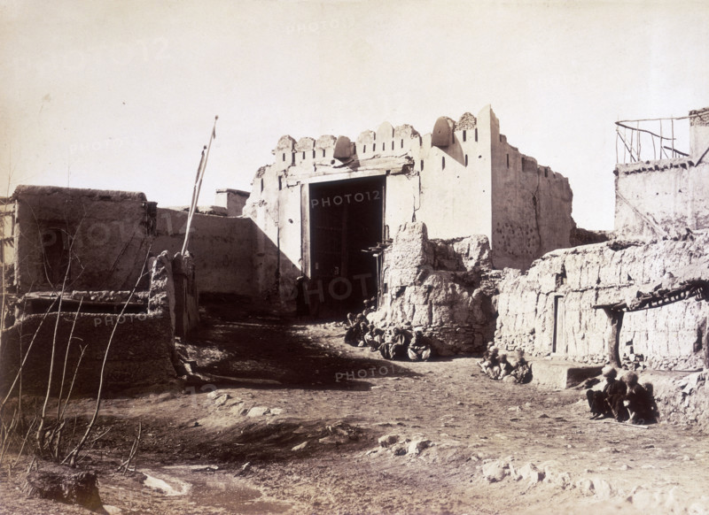 Lahore Gate, Kabul, Afghanistan, c1879. Small groups of people sit along the wall. A dirt track leads up to the gate which is in a delapidated state and falling into ruin.