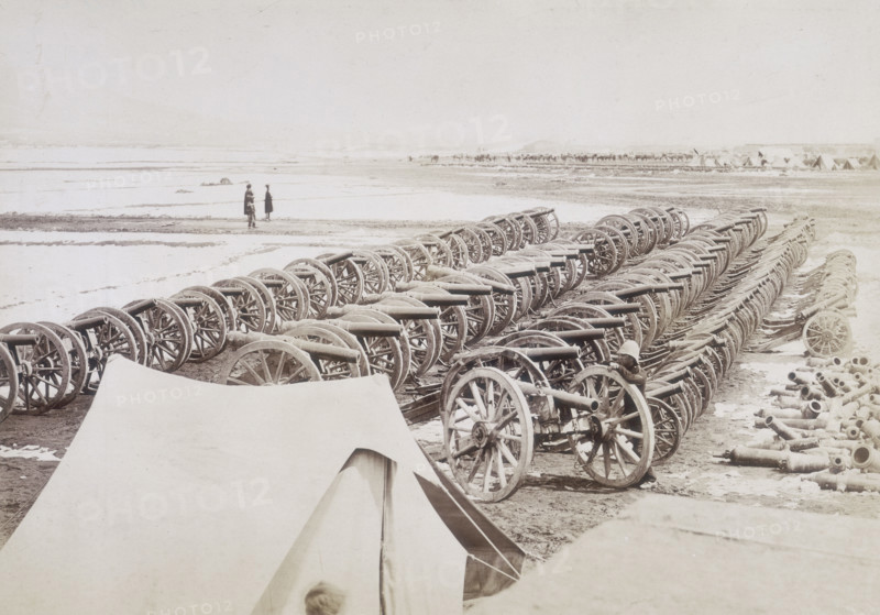 Captured guns, Kabul, Afghanistan, c1879. Cannons lined up in neat rows. A British soldier leans against a wheel for scale. On the right is a pile of dismantled guns. A camp can just be seen in the distance on the right.