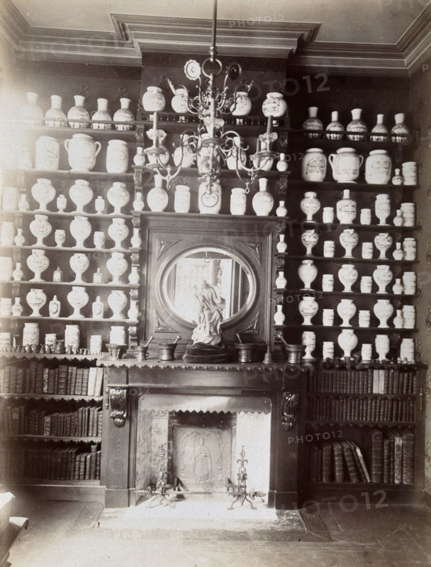 Drug jars and books on shelves surrounding a mantlepiece with a statue and a mirror, c1900.. Creator: Unknown.
