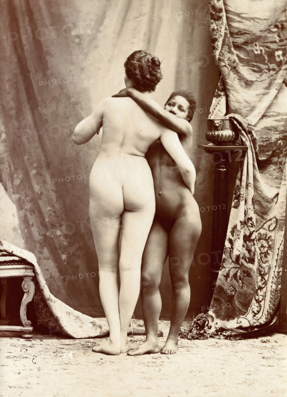 Two women posing naked in a photographic studio, standing embracing each other in front..., c1900. Creator: Unknown.