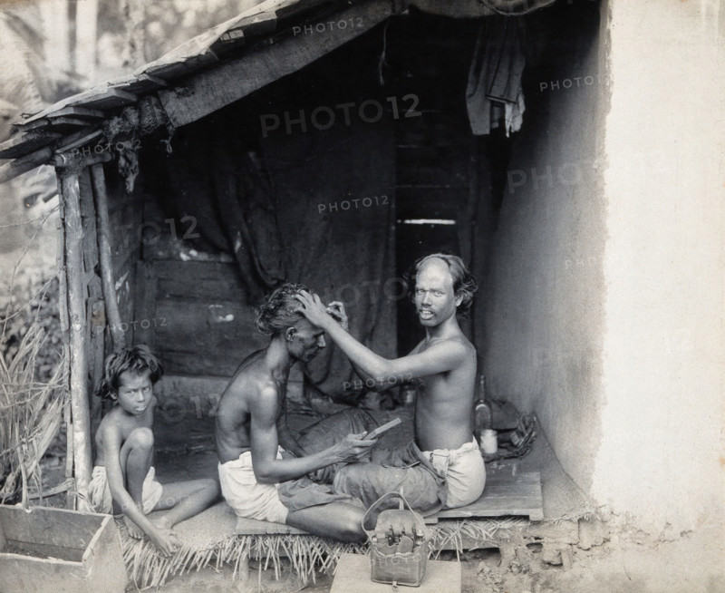 Two men sitting cross-legged; one is a barber shaving the front of the other's head, c1900. Creator: Unknown.