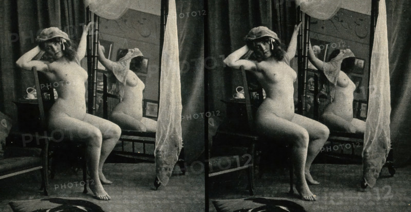 A woman posing naked in a photographic studio, sitting on a chair in front of a looking-glass, c1900 Creator: Unknown.
