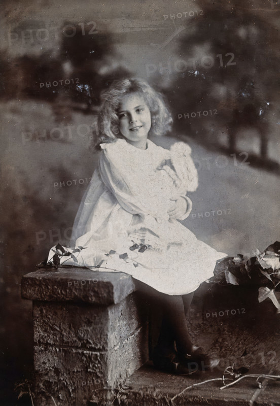 A girl, holding a doll, sitting on a wall, c1890s. Creator: Lock & Whitfield.
