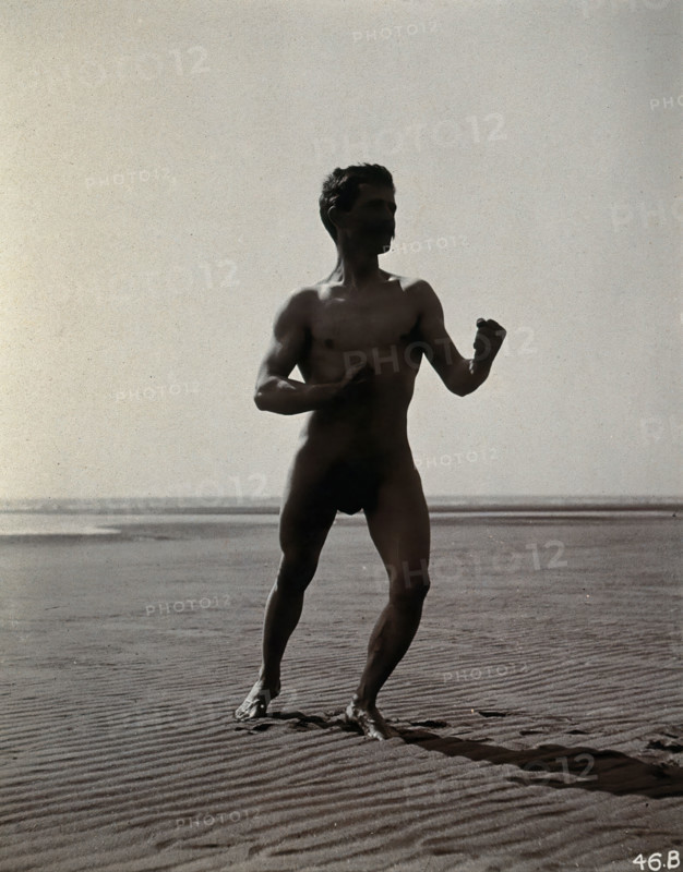 A man posing naked in a boxing stance with his fists raised, on a deserted beach, c1900. Creator: Unknown.