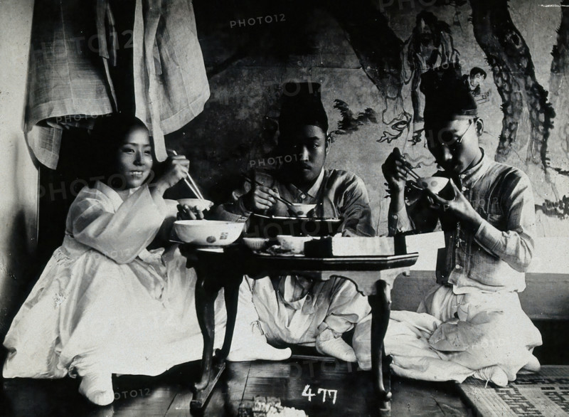 A group of people eating a meal, seated behind a small table, in front of a painted screen, c1900. Creator: Unknown.