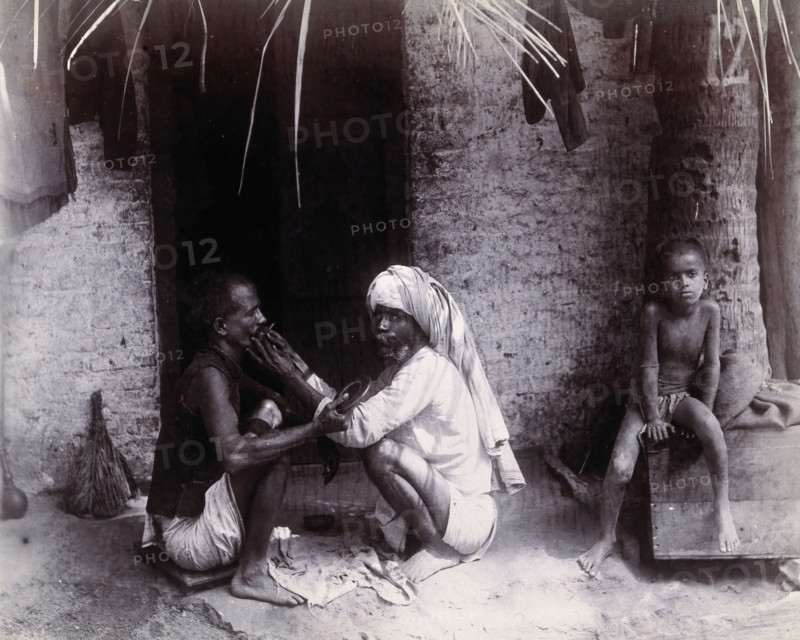 Two Indian men crouching; one is cutting the other's hair, while a girl sits.., c1890s.  Creator: Unknown.