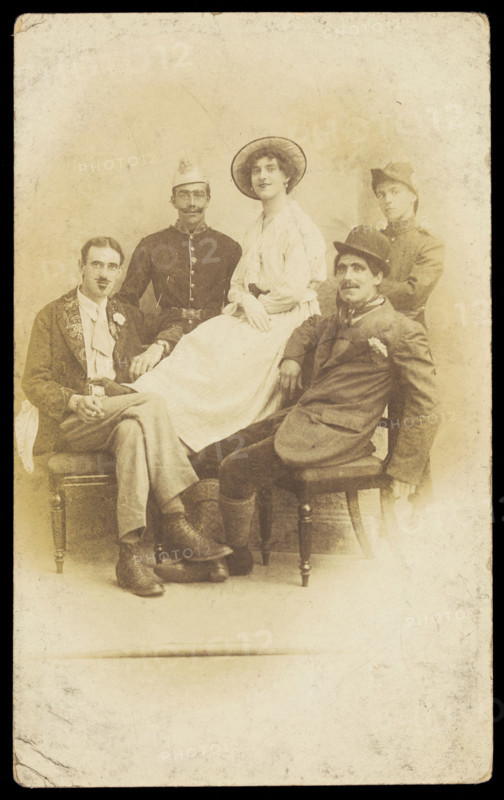 Amateur actors sitting down, surrounding a man in drag seated in the centre, [1910-1919?]. Creator: Unknown.