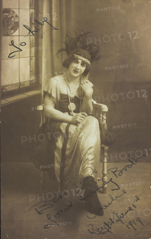 A man in drag (Charlie) sits face-on, wearing detailed costume, with a window painted..., 1919. Creator: Unknown.