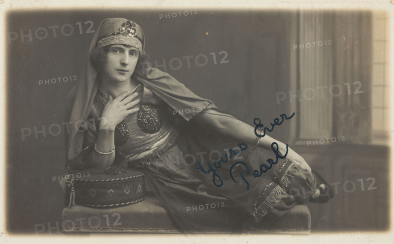 A man in drag, poses wearing delicate attire; curled up on a piece of furniture, [1910-1919?]. Creator: Unknown.