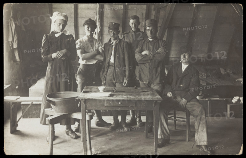 French prisoners of war, one in drag, gather round a small wooden table, [between 1910 and 1919?]. Creator: Unknown.