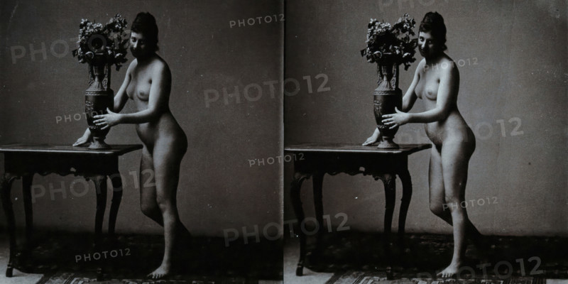 A young woman posing naked in a photographic studio, standing in front of a table on which..., c1900 Creator: Unknown.