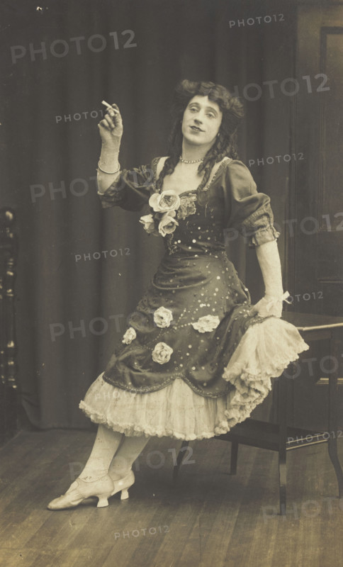 A performer in drag, wearing a large flowery dress, poses with a cigarette on stage, c1908-1910. Creator: Unknown.