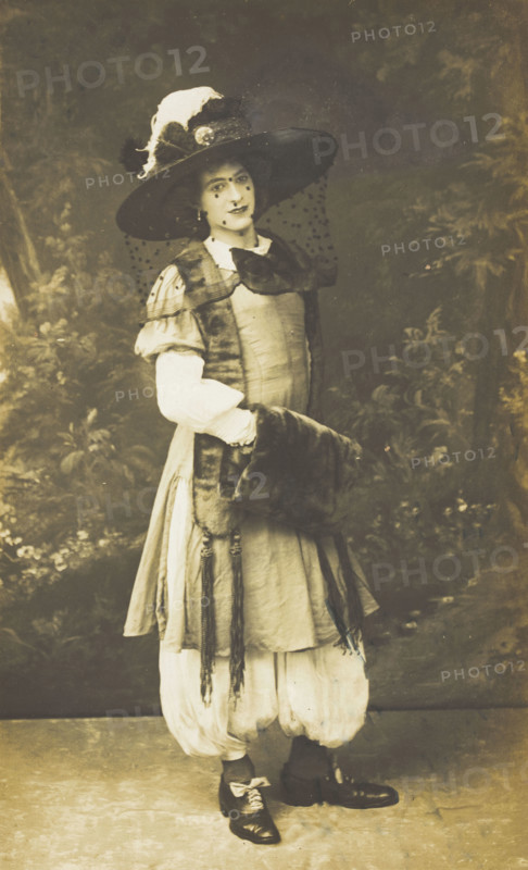 A man in drag wearing a very large hat with a veil, stands in front of a painted..., 1911. Creator: Unknown.