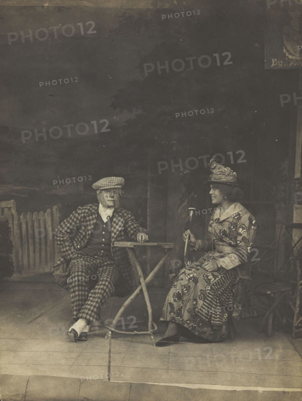 Two sailors, one in drag, sit on stage at a table, in front of very detailed scenery (1910-1919?). Creator: Unknown.