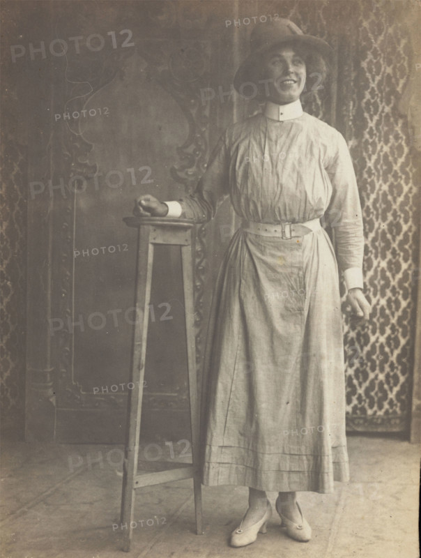 A sailor in drag leans on a high stool, in front of patterned scenery, (between 1910 and 1919?). Creator: Unknown.