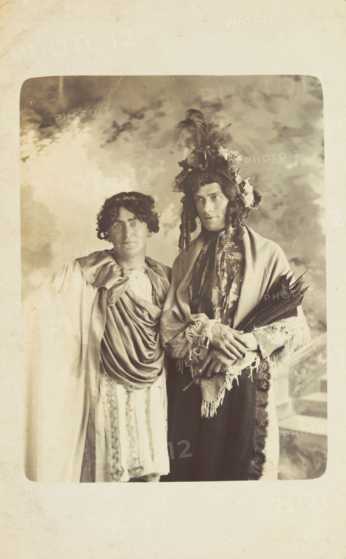 Two men in drag, one wearing a detailed feathered head garment, pose for a portrait, [1900-1909?]. Creator: Frank Wood.
