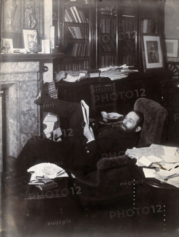 Arthur Schuster, lying on an armchair by a fireplace, with a photograph of Lord Kelvin..., c1901. Creator: Unknown.