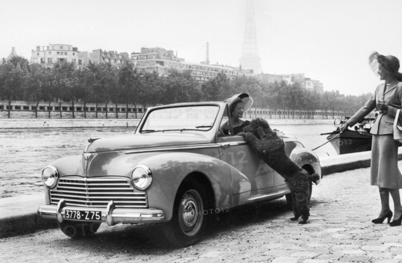 En octobre 1951 sur les berges de la Seine à Paris, à l'occasion du Salon de l'automobile, d'élégantes jeunes femmes présentent le nouveau modèle de la marque PEUGEOT, la 203. Née dans les ateliers de La Garenne en région parisienne, la 203 possède 42,5 chevaux et atteint une vitesse de 110/115 km/h. Différents millésimes seront produits jusqu'en 1956. A l'arrière plan, on distingue la silhouette embrumée de la tour Eiffel.