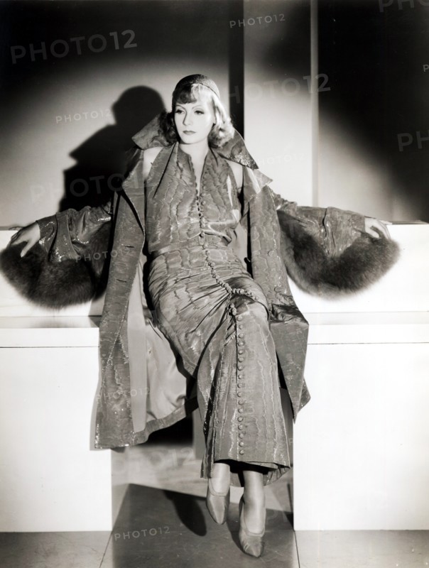 Dans les années 1930, l'actrice Greta Garbo habillée par Adrian, dessinateur de mode de la Metropolitan GoldWyn Mayer. Elle porte une robe de tissu vert sombre, broché d'argent et d'un manteau de la même couleur.   Greta Garbo (1905 - Stockholm / 1990 - New York). Modèle pour des publicités en Suède, Greta Garbo est remarquée par le réalisateur Eric Petscher. Il lui donne un petit rôle dans "Peter le Vagabond". Elle prend alors des cours de théâtre à l'Académie royale de Stockholm. Séduit, Mauritz Stiller lui confie un rôle majeur dans "La Légende". Garbo l'accompagne aux Etats-Unis. Elle est propulsée en tête d'affiche des films du cinéma muet, "Le Torrent", "La Chair et le Diable", "Anna Karénine". Malgré la fin de ce cinéma, sa carrière prend un nouveau tournant. Le public découvre sa voix dans "Anna Christie" en 1930. Elle enchaîne ensuite des rôles de séductrice dans "Mata Hari", "Le roman de Marguerite Gautier" et le plus dramatique "Ninotchka". Adulée, elle reste secrète et loin...