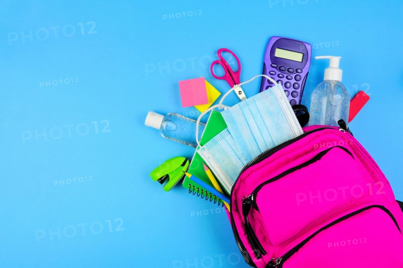 Backpack full of school supplies and COVID 19 prevention items. Top view, spilling onto a blue background. Back to school during pandemic concept.