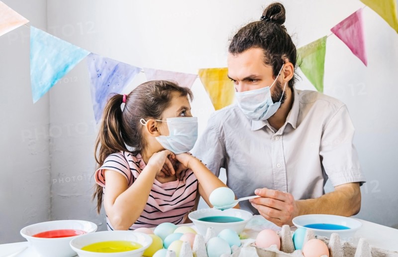 New normal Easter celebration lifestyle concept. Cute little girl and her dad wearing protective masks, preparing easter, dyed eggs at home during