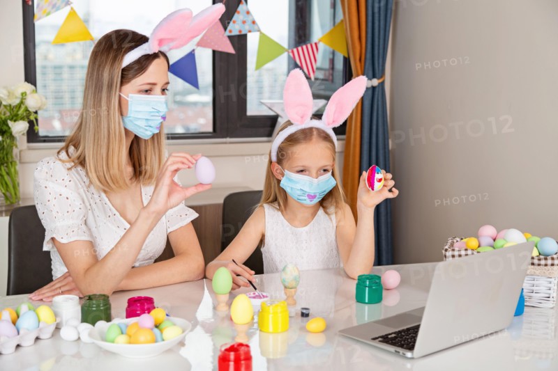 Pretty little girl wearing bunny ears and disposable medical face mask and painting easter eggs at home during coronavirus covid-19 outbreak.