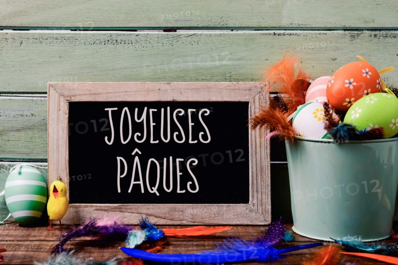 a chalkboard with the text joyeuses paques, happy easter in French, hanging on a rustic wooden wall, and a pile of different decorated easter eggs, a
