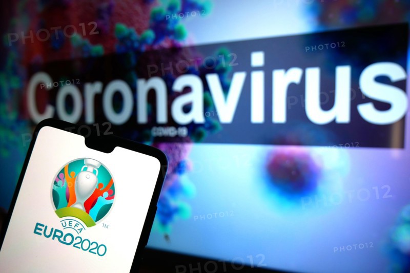 The UEFA Euro 2020 logo seen displayed on a mobile phone with an illustrative model of the Coronavirus displayed on a monitor in the background.