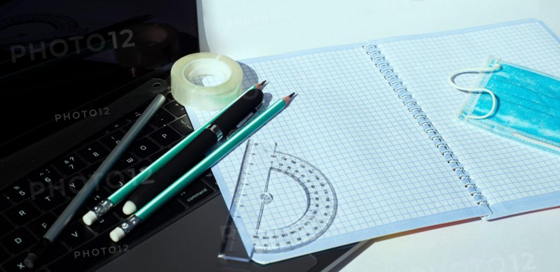 On the laptop keyboard lie a notebook pencils pens ball-point protractor adhesive tape and a disposable medical mask.