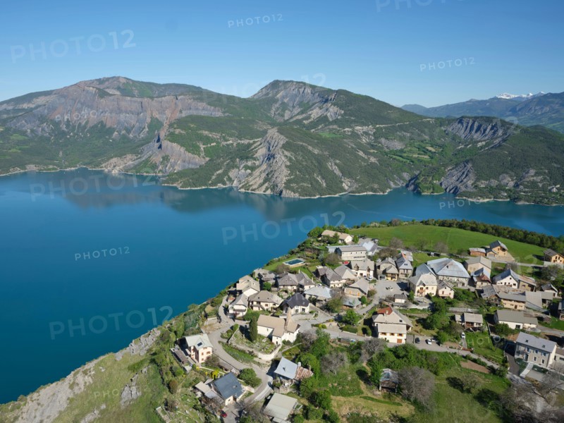 AERIAL VIEW - Village perched 250m-high above Lake Serre-Poncon. Le Sauze-du-Lac, Hautes Alpes, France.