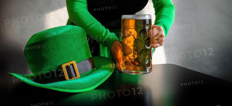 Woman's hand holding glass of beer near St. Patrick's day hat of a leprechaun on the black background