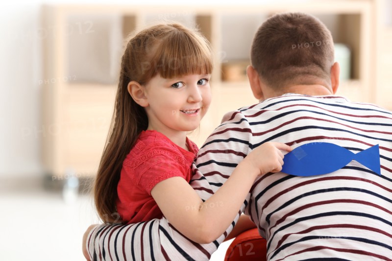 Little girl sticking paper fish to her father's back indoors. April fool's day prank
