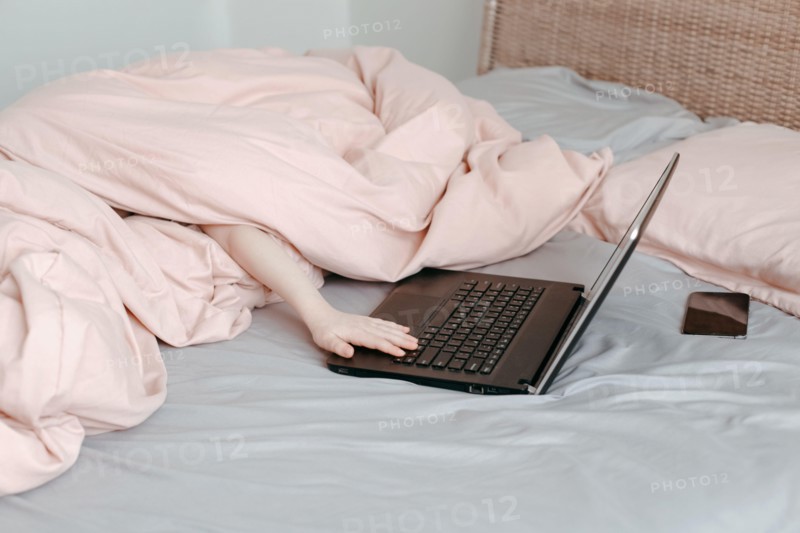 Bored funny child lying in bed under blanket and learning in virtual online school class. Hands typing on computer keyboard. Kid working on laptop