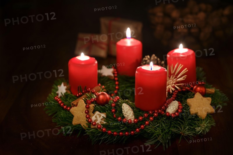 Advent wreath from fir branches with red lit candles and Christmas decoration, some gifts blurred in the dark background, copy space, selected focus,