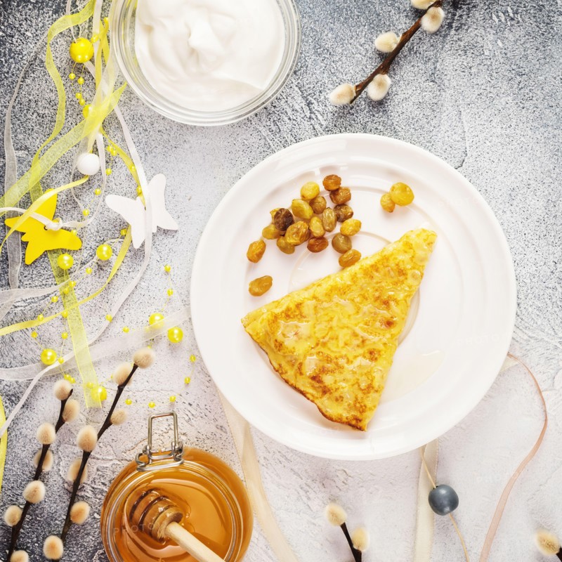 Sweet homemade fried crepe with honey, raisins on a white plate with holiday decoration, sour cream and honey on a blue background. Russian national f