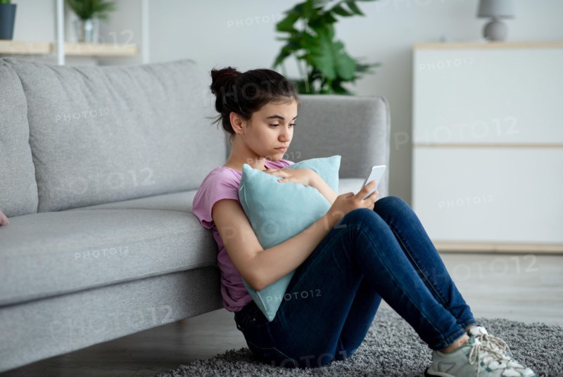 Stressed Indian teen girl sitting on floor with cellphone and reading bad message at home