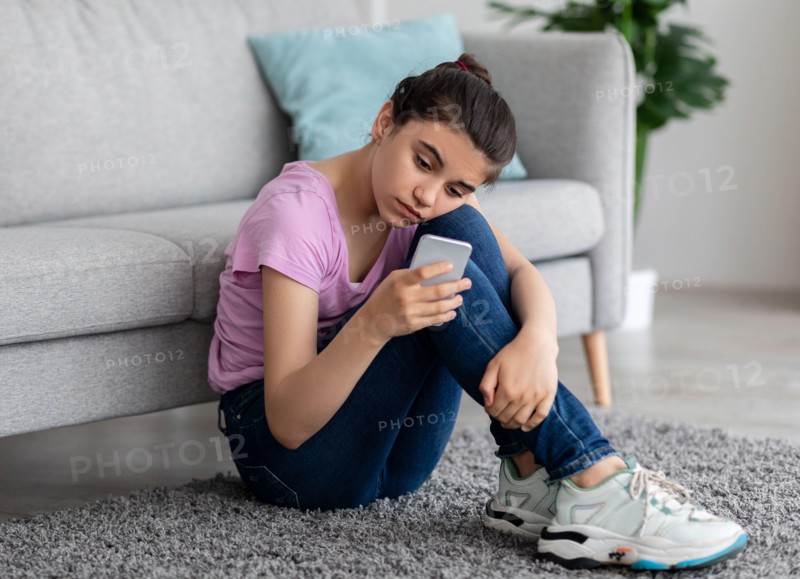 Unhappy Indian teen girl with mobile phone feeling sad, suffering from cyber bullying online, sitting on floor at home