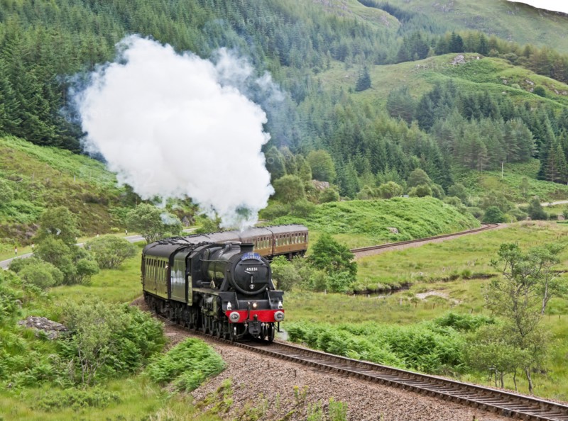 Jacobite steam train moves up hill beyond Glenfinnan Station towards Mallaig on a sunny summer day