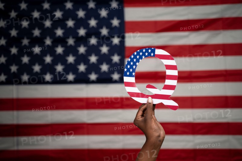 Hands holding Q alphabet with US flag as background with copy space - Concept QAnon or Q Anon deep state conspiracy theory